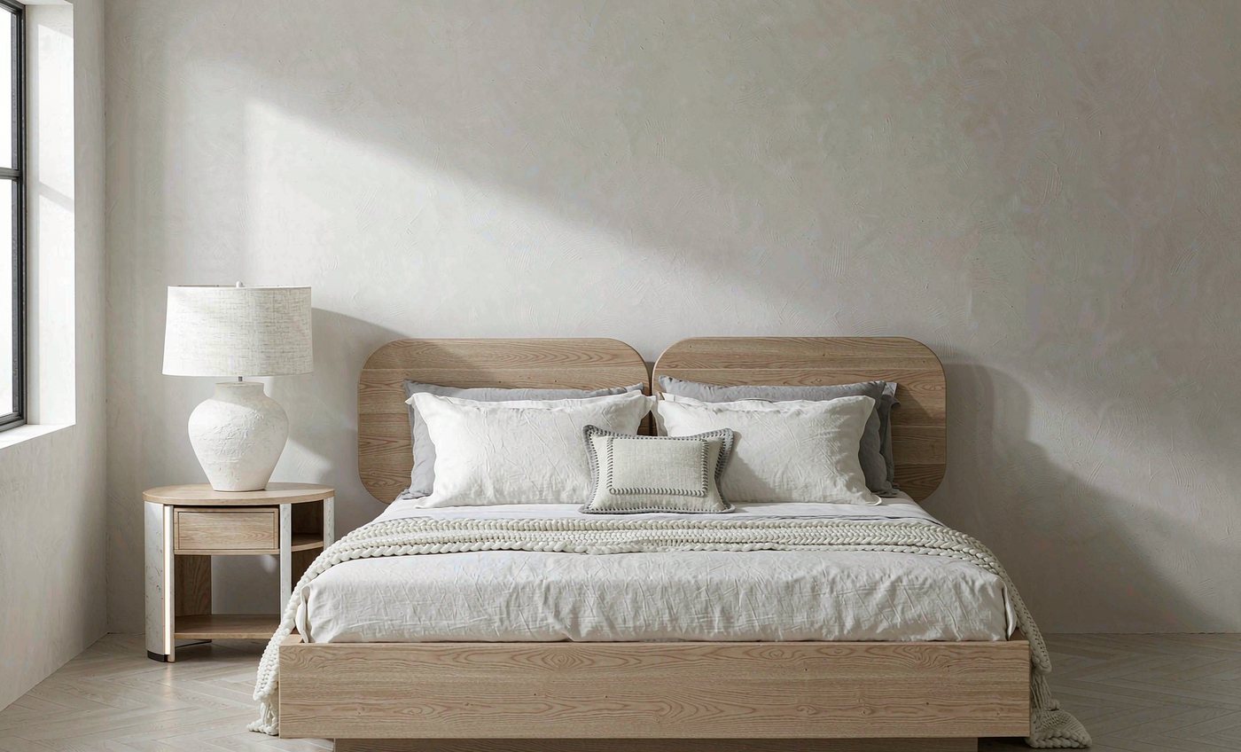 Low oak platform bed with layered white linen bedding and a nightstand lamp